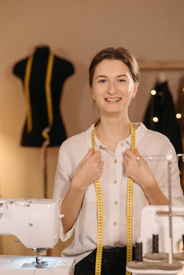 A happy female tailor in a sewing studio with measuring tape around her neck and sewing machines in view.